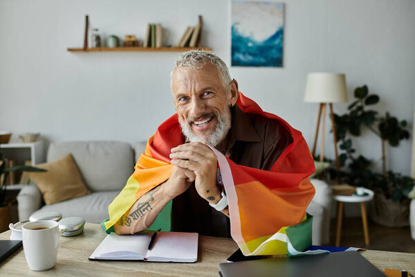 Взрослый гей с татуировками и седыми волосами гордо улыбается, держа в гостиной радужный флаг.