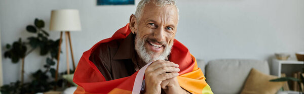 A mature, tattooed gay man with grey hair smiles while holding a pride rainbow flag at home.