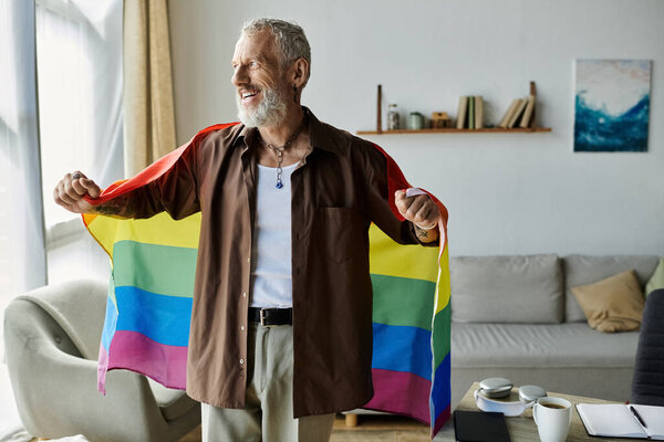 A mature gay man with tattoos and gray hair holds a rainbow pride flag at home, smiling.