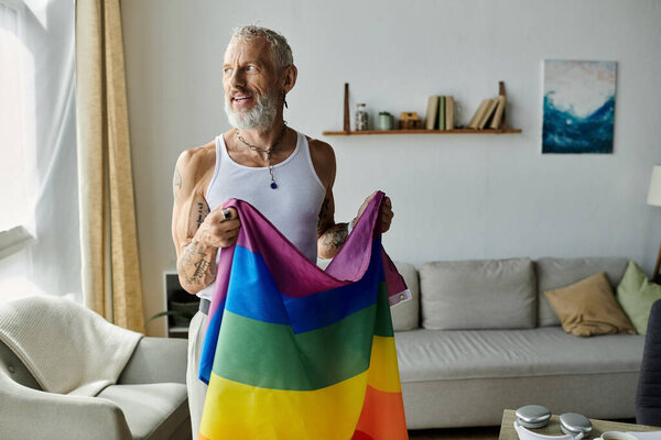 A mature gay man with tattoos and grey hair smiles and holds a pride rainbow flag in his living room.