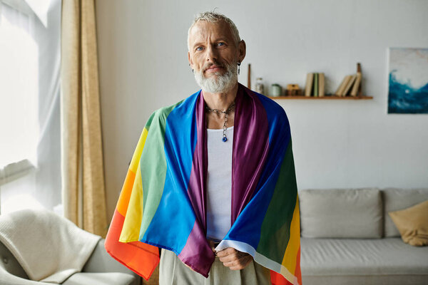 A mature gay man with tattoos and gray hair stands in his home, proudly holding a rainbow pride flag.