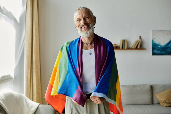 A mature gay man with grey hair and tattoos smiles while holding a rainbow pride flag at home.