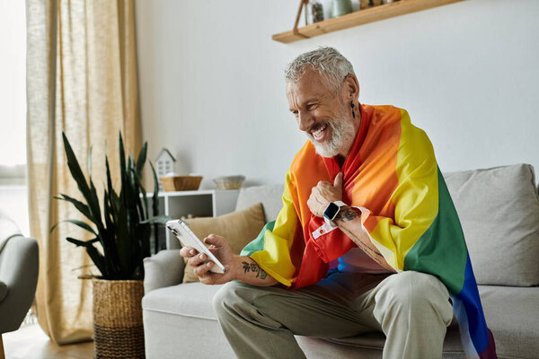 A mature gay man with grey hair and tattoos smiles while holding a pride flag. He is sitting on a couch in a home setting.