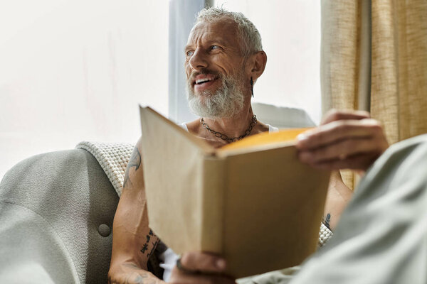 Татуированный пожилой гей с седой бородой расслабляется дома на диване, погружаясь в мысли во время чтения книги..