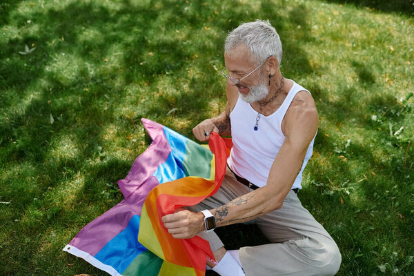 A mature gay man with tattoos and a grey beard sits on the grass holding a rainbow flag.