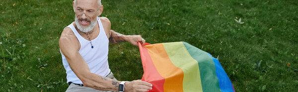 A mature gay man with tattoos and a grey beard holds a rainbow flag outdoors.