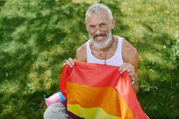 A mature gay man with a grey beard and tattoos smiles as he holds a rainbow flag outdoors.