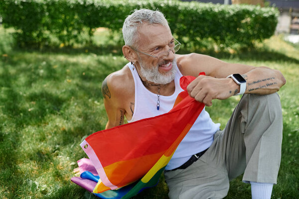 A mature gay man with tattoos and a grey beard holds a rainbow flag in a park. He smiles as he sits on the grass.
