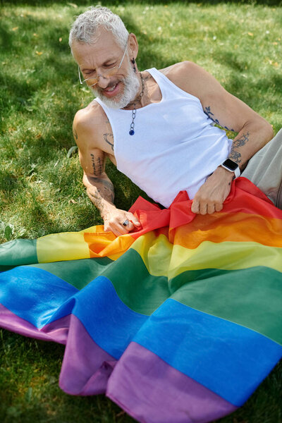 A mature man with tattoos and a grey beard smiles while holding a rainbow flag outdoors.