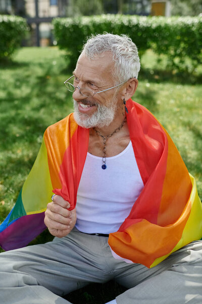 A mature gay man with tattoos and a grey beard smiles while holding a rainbow flag outdoors.