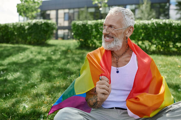 A mature gay man with tattoos and a grey beard smiles as he holds a rainbow flag in a park.