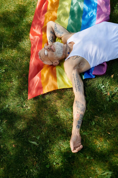A mature gay man with tattoos and a grey beard rests on a rainbow flag in a park, enjoying a sunny day.