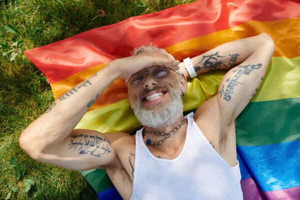A mature gay man with tattoos and a grey beard smiles while lying on a rainbow flag outdoors.