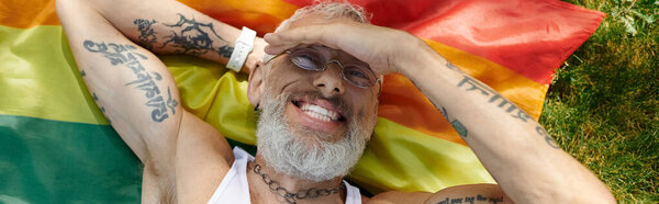 A mature gay man with tattoos and a grey beard lies on a rainbow flag outdoors, smiling.
