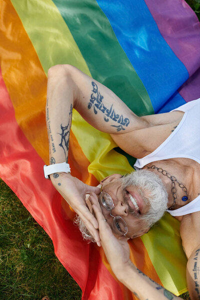 A mature gay man with tattoos and a grey beard smiles while holding a rainbow flag outdoors.
