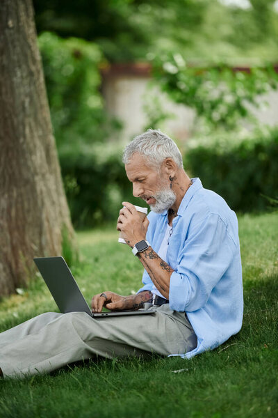 A man with tattoos and a grey beard sits on the grass, drinking coffee while working on his laptop.