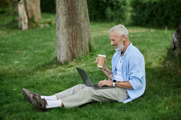 A mature man with tattoos and a grey beard sits on the grass in a park, working on his laptop while holding a coffee cup.