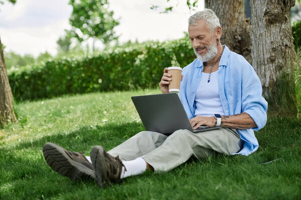 A mature gay man with tattoos and a grey beard sits on the grass outdoors, enjoying a coffee while working on his laptop.