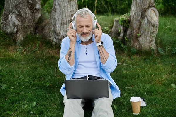 A mature gay man with tattoos and a grey beard sits on the grass in a park, enjoying music through headphones while using his laptop.