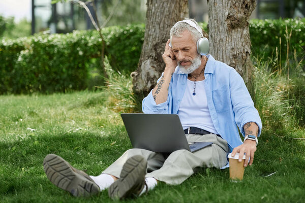 A mature gay man with tattoos and a grey beard sits on the grass outdoors, listening to music on headphones while using a laptop.