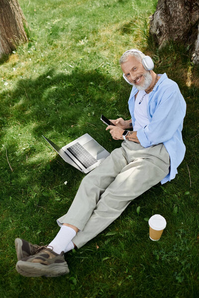 A mature gay man with tattoos and a grey beard sits on the grass, smiling, with a laptop and phone in his lap, wearing headphones.