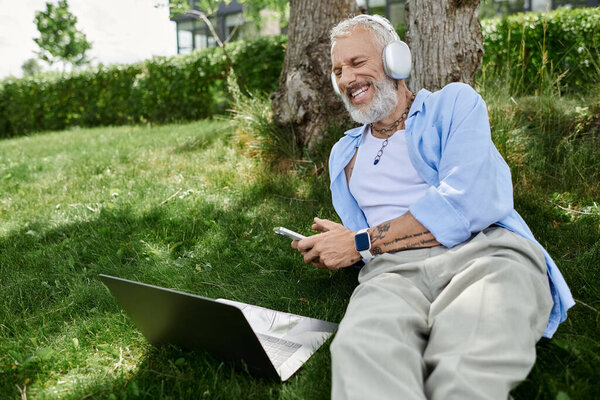 A mature man with tattoos and a grey beard enjoys a day outdoors, listening to music with headphones and working on his laptop.