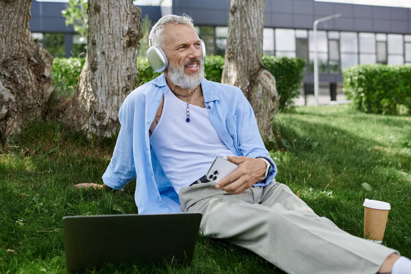 A mature gay man with tattoos and a grey beard sits on the grass in a park, enjoying the outdoors and listening to music on his headphones.