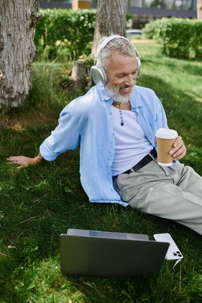 A mature man with tattoos and a grey beard sits on the grass in a park, enjoying a cup of coffee and listening to music with headphones.
