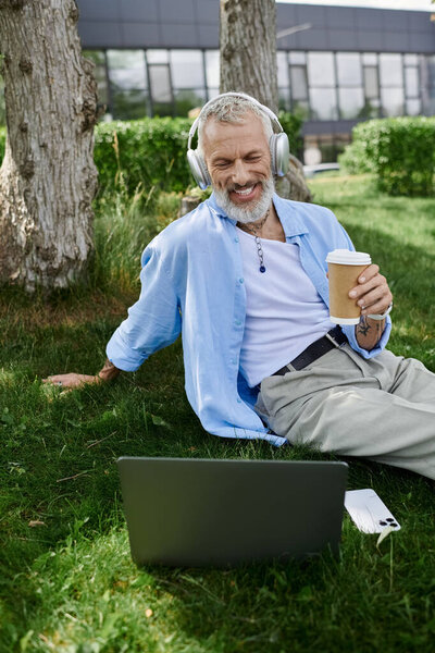 A mature man with tattoos and a grey beard sits on the grass outside, enjoying a coffee break with headphones on and a laptop open.