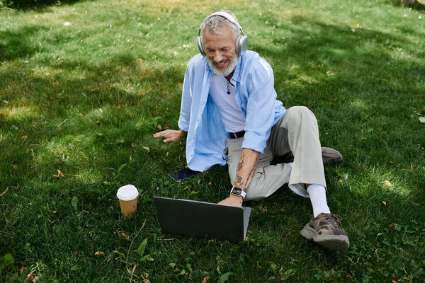 A mature gay man with tattoos and a grey beard is sitting on the grass in a park, working on his laptop.