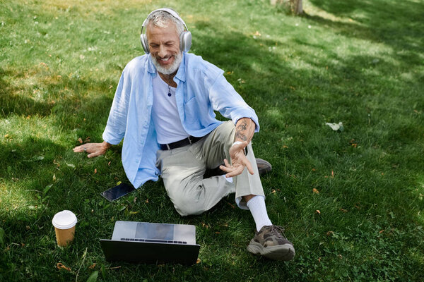A mature gay man with tattoos and a gray beard sits on the grass outdoors, wearing headphones and smiling.