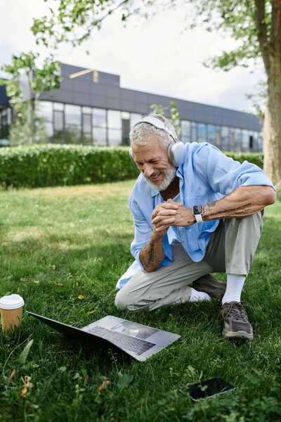A mature man with tattoos and a grey beard sits on the grass in a park wearing headphones and using a laptop.