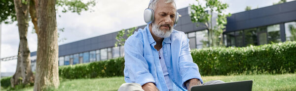 A mature gay man with tattoos and a grey beard sits on the grass outdoors, wearing headphones and listening to music.