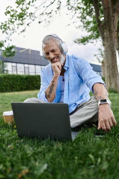 Man with tattoos and grey beard sitting on grass in park, working on laptop, wearing headphones, and looking relaxed and focused.