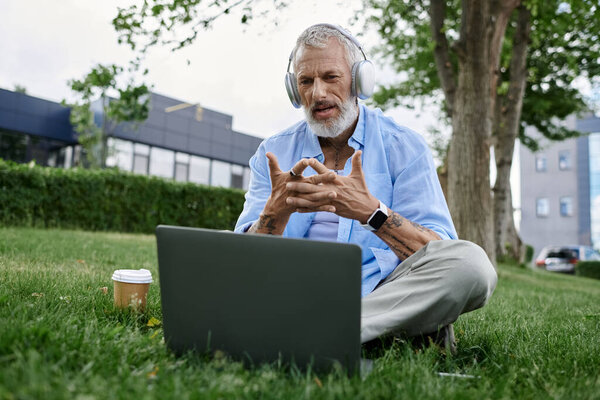 Tattooed man with grey beard sits on park grass, working on laptop with coffee cup and headphones.