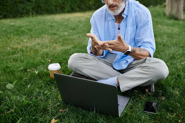 A mature gay man with tattoos and a grey beard sits on the grass outdoors, using a laptop.