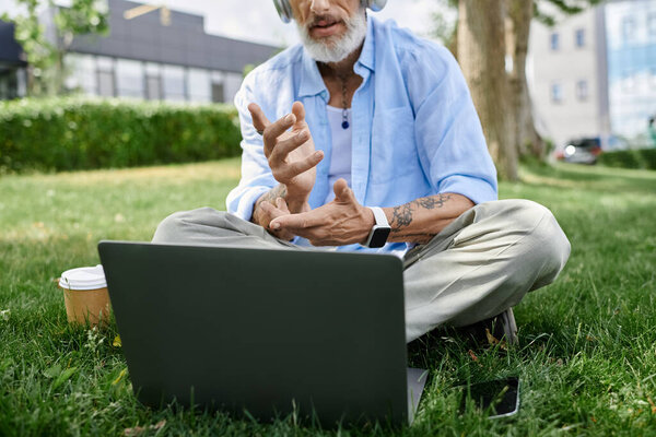 A mature gay man with tattoos and a grey beard sits on the grass outdoors, working on a laptop.