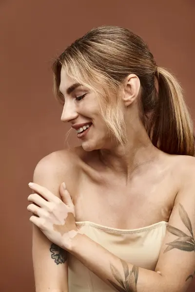 stock image A young woman with vitiligo and a tattoo smiles while posing with her shoulders exposed against a beige background.