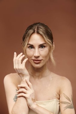 Vitiligo 'su ve dövmesi olan genç bir kadın bej arka planda çıplak omuzlarla poz veriyor..