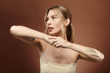 Vitiligo 'su olan ve kolunda dövmesi olan genç bir kadın omuzları bej arka planda çıplak poz veriyor..