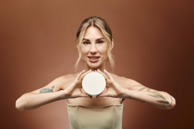 Vitiligo 'su olan genç bir kadın bej arka planda krema kavanozu tutuyor. Gülümsüyor ve kameraya bakıyor..