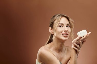 Vitiligo 'su olan genç bir kadın, bej arka planda bir kavanoz krema tutarken özgüvenle gülümsüyor..