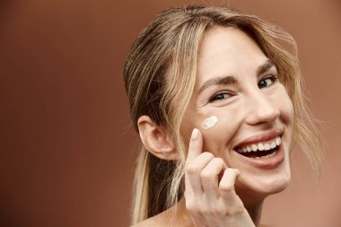 Vitiligo 'su olan genç bir kadın yüzüne krem sürüyor, parlak bir şekilde bej arka plana gülümsüyor..
