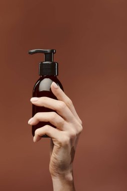 A young girls hand holds a dark brown skincare bottle against a beige background. clipart