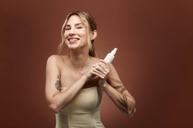 Vitiligo 'su olan genç bir kadın, bej bir arka plan önünde elinde bir cilt ürünü tutarken özgüvenle gülümsüyor..