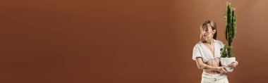 Vitiligo 'su olan genç bir kadın, bej bir arka plana karşı beyaz bir yaz elbisesi içinde, elinde saksıda kaktüs tutarak güven içinde poz veriyor..