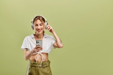 Vitiligo kulaklıklı bir kadın yeşil arka planda gülümsüyor, beyaz gömlek ve haki pantolon giyiyor, telefonunu kullanıyor..