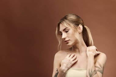 Vitiligo 'su olan ve kolunda dövmesi olan genç bir kadın bej arka planda çıplak omuzlarla poz veriyor..