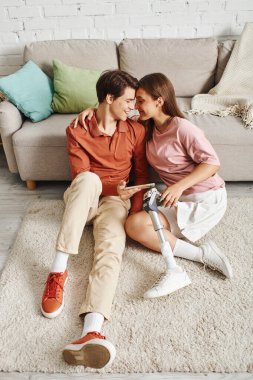 Couple on fluffy carpet shares tender moment; woman with prosthetic leg holds boyfriends hand. clipart