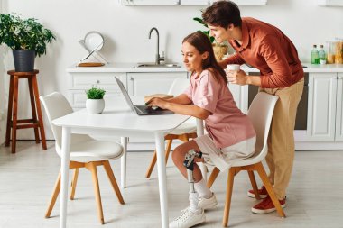 Erkek arkadaşı mutfakta beklerken protez bacaklı bir kadın dizüstü bilgisayarın başında duruyor..